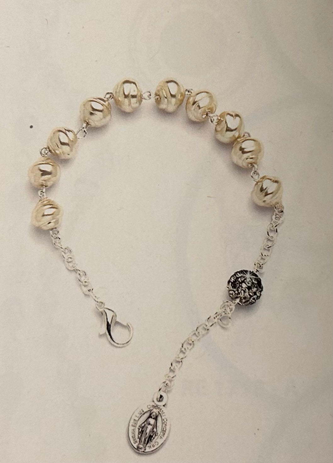 A silver-toned beaded bracelet featuring metallic pearl-like beads, a black-and-silver patterned spacer bead, and a dangling Miraculous Medal charm on a light beige background.