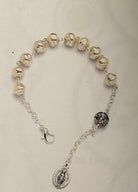 A silver-toned beaded bracelet featuring metallic pearl-like beads, a black-and-silver patterned spacer bead, and a dangling Miraculous Medal charm on a light beige background.